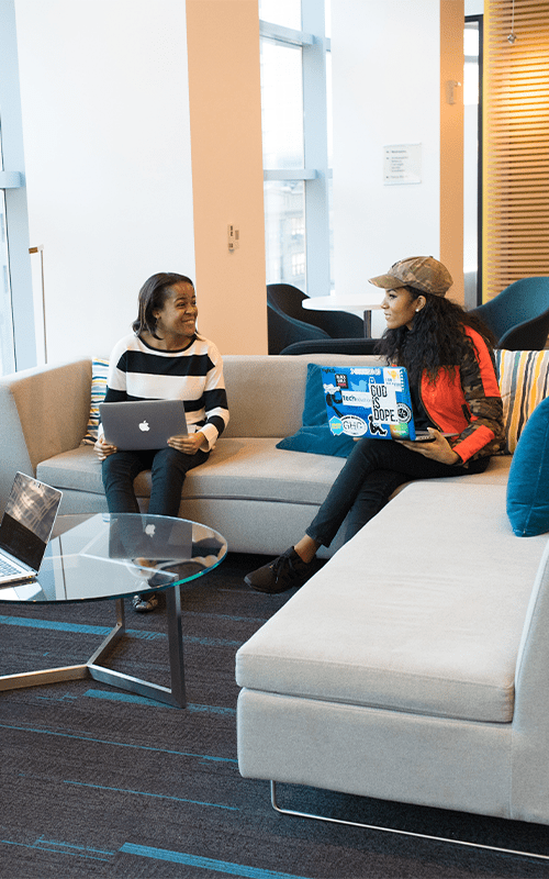 Two women sitting on a couch with laptops.